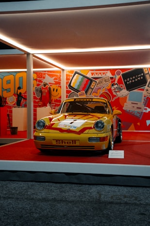 A vintage yellow sports car with red accents is displayed on a stage surrounded by vibrant 90s-themed decor. The backdrop features colorful graphics like a retro television, sneakers, a smiley face, and a basketball jersey, creating a nostalgic atmosphere.
