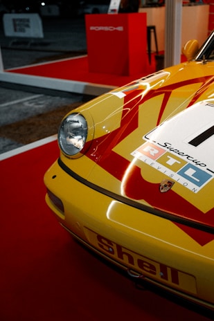 A close-up of a vintage yellow Porsche car featuring red racing stripes and a prominent headlight. The car has racing stickers, including 'RTL' and a Porsche emblem. It is displayed on a red carpet in a showroom environment, with Porsche branding visible in the background.