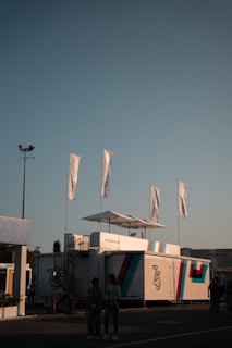 A modern exhibition setup with a sleek white structure displaying the number 75 and the Porsche logo. Five tall white flags flutter above. Two people stand in the foreground, partially illuminated by soft lighting, with a clear evening sky providing background contrast.