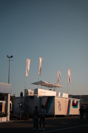 A modern exhibition setup with a sleek white structure displaying the number 75 and the Porsche logo. Five tall white flags flutter above. Two people stand in the foreground, partially illuminated by soft lighting, with a clear evening sky providing background contrast.