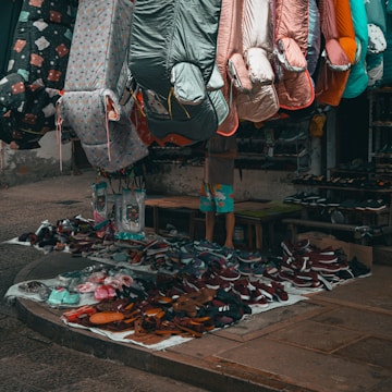 A street market stall is filled with colorful sleeping bags hanging above a variety of shoes neatly arranged on the ground. The sleeping bags feature different colors and patterns, and the footwear includes sandals and sneakers. A person is partially visible behind the stall, wearing shorts with bright patterns.