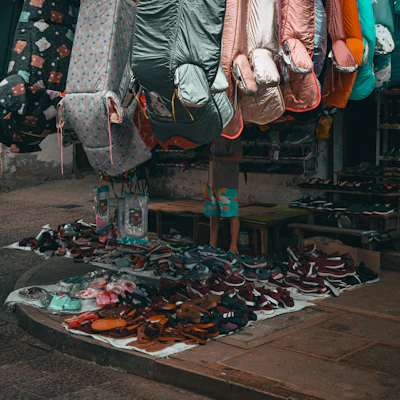 A street market stall is filled with colorful sleeping bags hanging above a variety of shoes neatly arranged on the ground. The sleeping bags feature different colors and patterns, and the footwear includes sandals and sneakers. A person is partially visible behind the stall, wearing shorts with bright patterns.