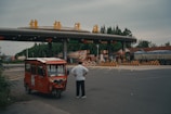 A man stands with his hands on his hips, observing a toll booth or entrance of a highway. An orange three-wheeled vehicle is parked nearby. The booth has yellow signage in Chinese and several lanes marked with road signs and barriers. Surrounding the area are trees and large vehicles.