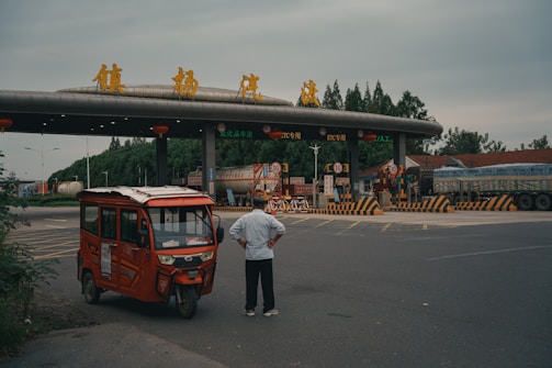 A man stands with his hands on his hips, observing a toll booth or entrance of a highway. An orange three-wheeled vehicle is parked nearby. The booth has yellow signage in Chinese and several lanes marked with road signs and barriers. Surrounding the area are trees and large vehicles.