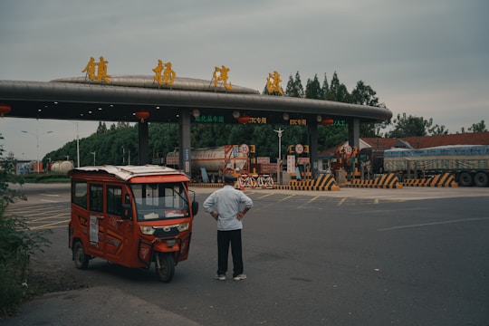 A man stands with his hands on his hips, observing a toll booth or entrance of a highway. An orange three-wheeled vehicle is parked nearby. The booth has yellow signage in Chinese and several lanes marked with road signs and barriers. Surrounding the area are trees and large vehicles.