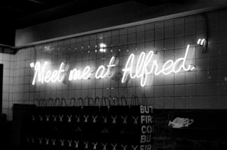 Neon sign displaying the phrase 'Meet me at Alfred' mounted on a tiled wall. Below the sign are several shopping bags with visible text and logos. The image has a monochromatic, grayscale color scheme that gives it a vintage feel.