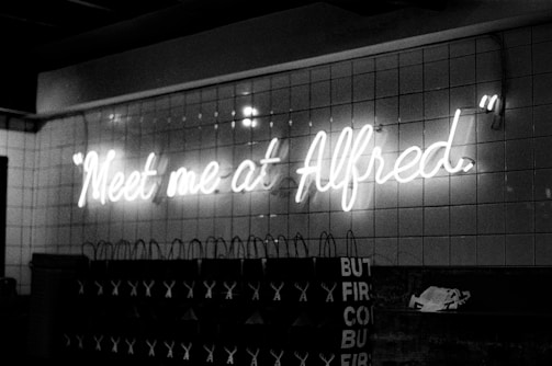 Neon sign displaying the phrase 'Meet me at Alfred' mounted on a tiled wall. Below the sign are several shopping bags with visible text and logos. The image has a monochromatic, grayscale color scheme that gives it a vintage feel.