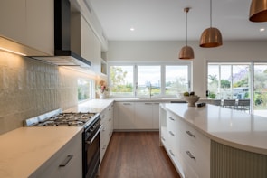 A modern kitchen features sleek white cabinetry and a large island with drawers and elegant curved edges. Stainless steel appliances, including a stove and extractor fan, are built into the cabinets. Large windows fill the space with natural light, and copper pendant lights hang from the ceiling. The kitchen opens to a patio with outdoor furniture, and lush greenery is visible through the windows.