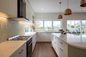A modern kitchen features sleek white cabinetry and a large island with drawers and elegant curved edges. Stainless steel appliances, including a stove and extractor fan, are built into the cabinets. Large windows fill the space with natural light, and copper pendant lights hang from the ceiling. The kitchen opens to a patio with outdoor furniture, and lush greenery is visible through the windows.