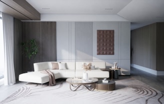 A modern, minimalist living room with a large white sectional sofa adorned with beige and brown pillows. A round coffee table is placed in front of the sofa, holding decorative items such as a vase with dried plants and a small bowl. On the wall behind the sofa, there is a modern art piece with a three-dimensional design. A large window with sheer curtains lets in natural light from the left side of the room. A tall green plant is placed in the corner between the window and the wall. The room is decorated in neutral colors with wooden and metallic accents.