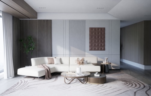A modern, minimalist living room with a large white sectional sofa adorned with beige and brown pillows. A round coffee table is placed in front of the sofa, holding decorative items such as a vase with dried plants and a small bowl. On the wall behind the sofa, there is a modern art piece with a three-dimensional design. A large window with sheer curtains lets in natural light from the left side of the room. A tall green plant is placed in the corner between the window and the wall. The room is decorated in neutral colors with wooden and metallic accents.