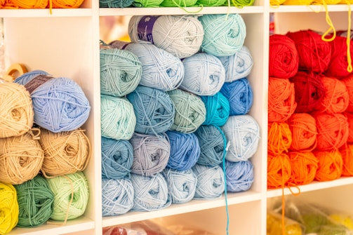 Shelves filled with neatly arranged yarn balls in various shades, including reds, oranges, blues, greens, and beiges. Each color is stacked in its own section, creating a vibrant and organized display.