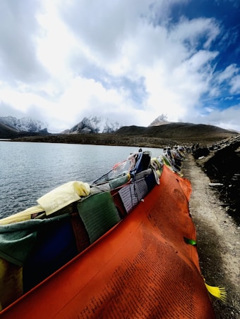 A serene mountain landscape featuring a lake surrounded by snow-capped peaks. In the foreground, colorful prayer flags are draped along a pathway beside the water, their vibrant hues contrasting with the muted tones of the natural surroundings. The sky is partly cloudy, adding depth to the scene.