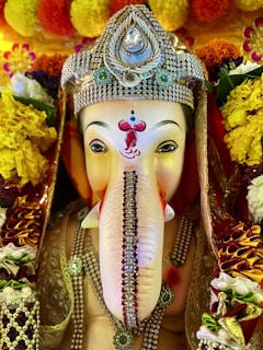 A beautifully adorned statue of the Hindu deity Ganesha, featuring intricate jewelry and vibrant floral decorations. The statue has a distinct, intricately decorated headdress with jewels and ornate patterns, while colorful flowers surround it, enhancing the deity's presence.