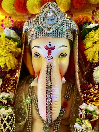 A beautifully adorned statue of the Hindu deity Ganesha, featuring intricate jewelry and vibrant floral decorations. The statue has a distinct, intricately decorated headdress with jewels and ornate patterns, while colorful flowers surround it, enhancing the deity's presence.