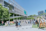 A lively outdoor area with groups of people walking and interacting near a modern building with a large screen displaying nature-themed images. Several market stalls or stands line the area, and lush greenery decorates parts of the building facade, adding a natural touch to the urban setting.