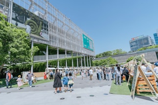 A lively outdoor area with groups of people walking and interacting near a modern building with a large screen displaying nature-themed images. Several market stalls or stands line the area, and lush greenery decorates parts of the building facade, adding a natural touch to the urban setting.