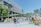 A lively outdoor area with groups of people walking and interacting near a modern building with a large screen displaying nature-themed images. Several market stalls or stands line the area, and lush greenery decorates parts of the building facade, adding a natural touch to the urban setting.