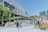 A lively outdoor area with groups of people walking and interacting near a modern building with a large screen displaying nature-themed images. Several market stalls or stands line the area, and lush greenery decorates parts of the building facade, adding a natural touch to the urban setting.