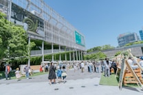 A lively outdoor area with groups of people walking and interacting near a modern building with a large screen displaying nature-themed images. Several market stalls or stands line the area, and lush greenery decorates parts of the building facade, adding a natural touch to the urban setting.