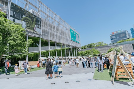 A lively outdoor area with groups of people walking and interacting near a modern building with a large screen displaying nature-themed images. Several market stalls or stands line the area, and lush greenery decorates parts of the building facade, adding a natural touch to the urban setting.