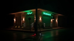 A dimly lit street corner featuring a closed pharmacy with bright green signage in Arabic and French. The storefront is illuminated by a series of warm lights, casting a subdued glow. A lone electric scooter is parked on the wet pavement under a streetlight.