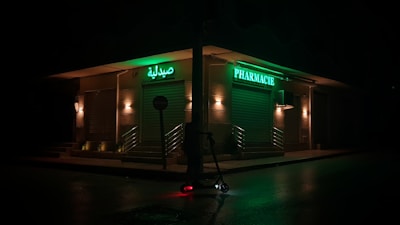 A dimly lit street corner featuring a closed pharmacy with bright green signage in Arabic and French. The storefront is illuminated by a series of warm lights, casting a subdued glow. A lone electric scooter is parked on the wet pavement under a streetlight.