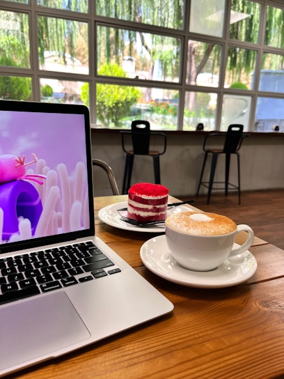 A cozy cafe setting with a laptop displaying an animated pink character on the screen. In front of the laptop sits a red and white layered cake on a plate and a cappuccino in a white cup and saucer. The background features a large window overlooking lush greenery and trees, with two black barstools positioned near the window.