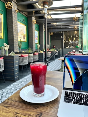 A modern cafe interior with green walls and geometric lighting fixtures. A refreshing red beverage in a tall glass sits on a white plate on a wooden table, next to an open laptop. The seating area features comfortable chairs and potted plants.