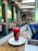 A modern cafe interior with green walls and geometric lighting fixtures. A refreshing red beverage in a tall glass sits on a white plate on a wooden table, next to an open laptop. The seating area features comfortable chairs and potted plants.