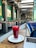 A modern cafe interior with green walls and geometric lighting fixtures. A refreshing red beverage in a tall glass sits on a white plate on a wooden table, next to an open laptop. The seating area features comfortable chairs and potted plants.