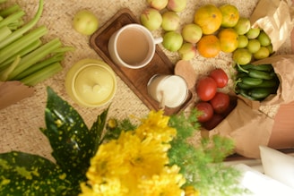 A variety of fruits and vegetables are arranged on a woven mat, including tomatoes, cucumbers, apples, oranges, and celery. A cup of beverage sits on a small wooden tray beside a glass jar with a wooden spoon. A yellow teapot and a bouquet of yellow flowers add vibrant colors to the scene.