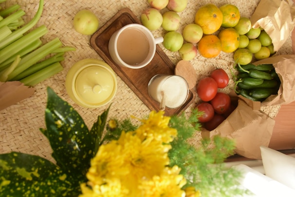 A variety of fruits and vegetables are arranged on a woven mat, including tomatoes, cucumbers, apples, oranges, and celery. A cup of beverage sits on a small wooden tray beside a glass jar with a wooden spoon. A yellow teapot and a bouquet of yellow flowers add vibrant colors to the scene.