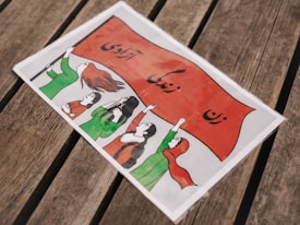 A group of women, illustrated in a cartoon style, are depicted holding up a large red banner. The women are all wearing clothes in shades of green and red. Their expressions and poses suggest a sense of determination and solidarity. The banner features text in a non-Latin script. The image appears on a wooden surface, adding a rustic texture to the background.