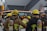 A group of firefighters in yellow helmets and protective gear gathered on a street intersection. They are equipped with oxygen tanks and are facing away from the camera, possibly preparing for or responding to an emergency. The background shows an urban setting with buildings and streetlights.