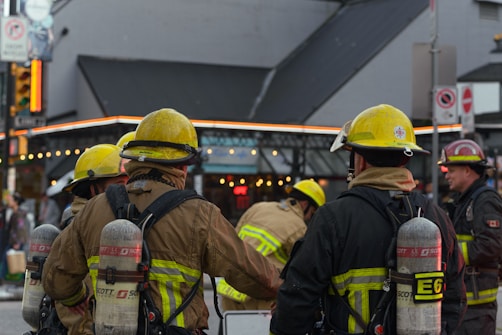 A group of firefighters in yellow helmets and protective gear gathered on a street intersection. They are equipped with oxygen tanks and are facing away from the camera, possibly preparing for or responding to an emergency. The background shows an urban setting with buildings and streetlights.