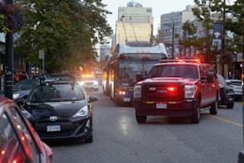A busy urban street scene featuring a red emergency vehicle with lights flashing, surrounded by other vehicles. A bus is prominently visible, and there are people walking on the sidewalks. Tall buildings and trees line the street in the background.