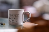 A ceramic mug with the words 'COFFEE fresh & authentic the best coffee in town' printed on it sits on a reflective surface. Nearby is a black and gold audio jack. The background is softly blurred, highlighting the mug as the focal point.