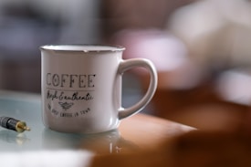 A ceramic mug with the words 'COFFEE fresh & authentic the best coffee in town' printed on it sits on a reflective surface. Nearby is a black and gold audio jack. The background is softly blurred, highlighting the mug as the focal point.