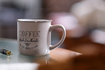 A ceramic mug with the words 'COFFEE fresh & authentic the best coffee in town' printed on it sits on a reflective surface. Nearby is a black and gold audio jack. The background is softly blurred, highlighting the mug as the focal point.