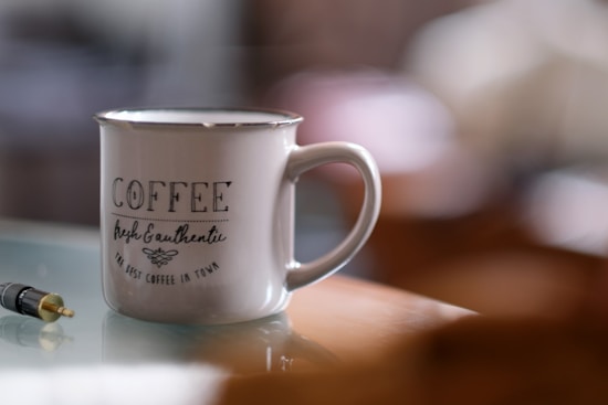 A ceramic mug with the words 'COFFEE fresh & authentic the best coffee in town' printed on it sits on a reflective surface. Nearby is a black and gold audio jack. The background is softly blurred, highlighting the mug as the focal point.