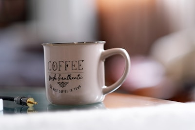 A ceramic mug with a large handle placed on a reflective surface, prominently featuring the text 'Coffee Fresh & Authentic The Best Coffee in Town'. The background is softly blurred, indicating a cozy indoor setting. A headphone jack is also visible on the surface near the mug.