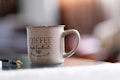 A ceramic mug with a large handle placed on a reflective surface, prominently featuring the text 'Coffee Fresh & Authentic The Best Coffee in Town'. The background is softly blurred, indicating a cozy indoor setting. A headphone jack is also visible on the surface near the mug.