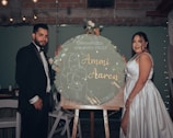 A couple is dressed in formal attire, standing beside a large circular sign on an easel. The man is wearing a black suit with a bow tie, while the woman is in a white satin dress. The sign features floral designs and the names 'Ammi Aaron' with a date. The background includes dim lighting and string lights, creating a celebratory atmosphere.