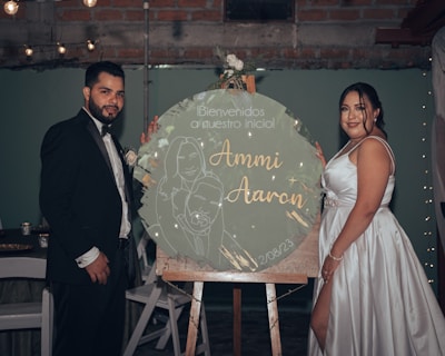 A couple is dressed in formal attire, standing beside a large circular sign on an easel. The man is wearing a black suit with a bow tie, while the woman is in a white satin dress. The sign features floral designs and the names 'Ammi Aaron' with a date. The background includes dim lighting and string lights, creating a celebratory atmosphere.