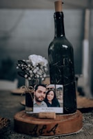 A large wine bottle with a cork sits on a wooden base that has engraved initials. Next to it, a small vase holds white flowers and decorative twine, accompanied by a printed photograph of a couple. The setup is on a textured table surface, suggesting a rustic or intimate setting.