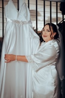A woman in a satin robe stands next to a hanging white wedding dress with a jeweled belt. She is smiling slightly and has her hand on the dress. There is a metal railing in the background.
