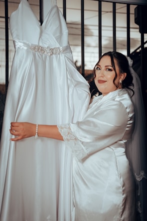 A woman in a satin robe stands next to a hanging white wedding dress with a jeweled belt. She is smiling slightly and has her hand on the dress. There is a metal railing in the background.