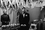 A formal gathering in a room decorated with cascading white flowers and draped fabric. A man in a suit stands beside a table adorned with floral arrangements, while another person in a patterned dress stands in the background, smiling. The ambiance is elegant and solemn.