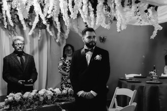 A formal gathering in a room decorated with cascading white flowers and draped fabric. A man in a suit stands beside a table adorned with floral arrangements, while another person in a patterned dress stands in the background, smiling. The ambiance is elegant and solemn.